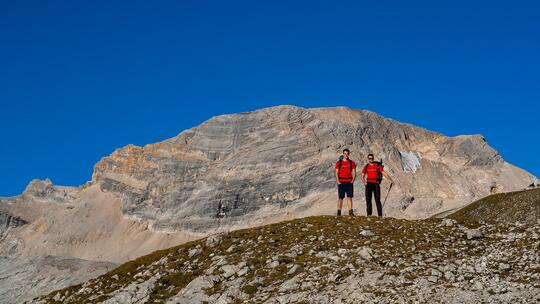 Two climbers from corpuls@Alpine stand on a hill in front of a mountain and blue sky
